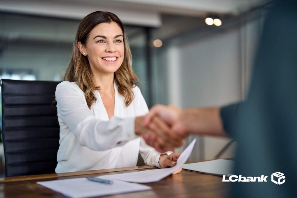Mulher sorrindo em um escritório enquanto aperta a mão de outra pessoa, segurando documentos sobre a mesa, representando um acordo concluído com o apoio do LCbank.